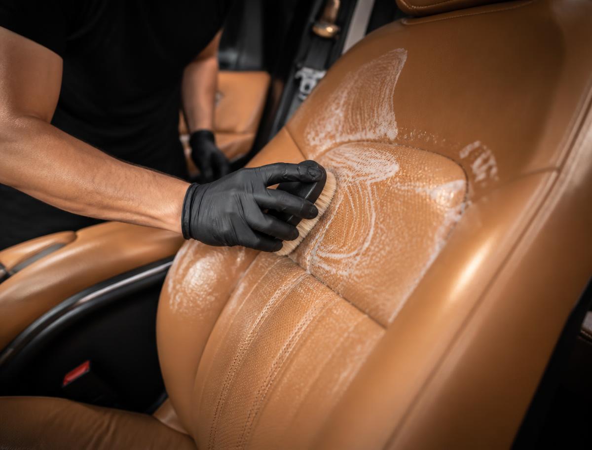Detailer cleaning a tan leather car seat with a soft brush and foam